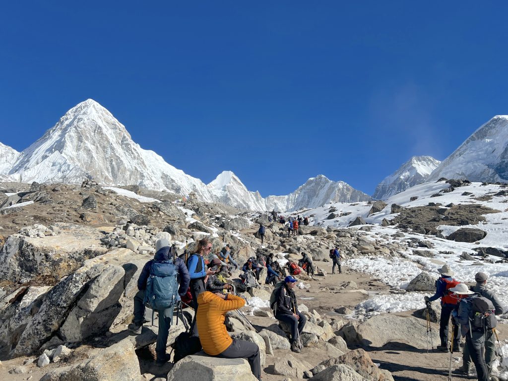 Wide shot of trekkers ascending a stone staircase above Namche Bazaar, prayer flags in the foreground, Everest visible in the distance